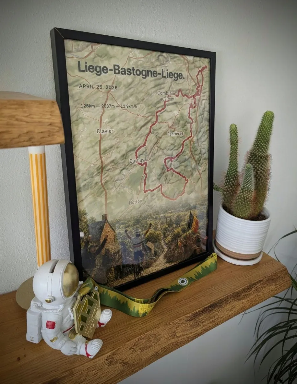 Pedros gerahmtes MementoMap-Poster der Liège-Bastogne-Liège auf einem Holzregal an einer weißen Wand, davor liegt seine Finisher-Medaille am grün-gelben Band des Rennens, gehalten von einer Astronautenfigur, rechts daneben ein Kaktus im weißen Topf, links eine gelb-weiß gestreifte Kerze